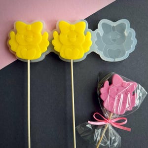 May include: Yellow chick-shaped lollipops on wooden sticks, with a bow on top, next to a silicone mold and a pink lollipop in a clear wrapper tied with a pink ribbon. The background is a mix of pink and black.