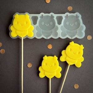 Può includere: Lecca lecca gialli a forma di ape su bastoncini di legno, con uno stampo in silicone trasparente sullo sfondo. Lo stampo ha tre forme di ape. Lo sfondo è grigio scuro con coriandoli marroni sparsi.