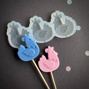 以下が含まれることがあります： 3つの鶏の形をしたシリコンモールドと、鶏の形をした2つのロリポップ。ロリポップは1つが青、もう1つがピンクで、どちらも木の棒に刺さっています。モールドは透明で、背景は濃いグレーです。