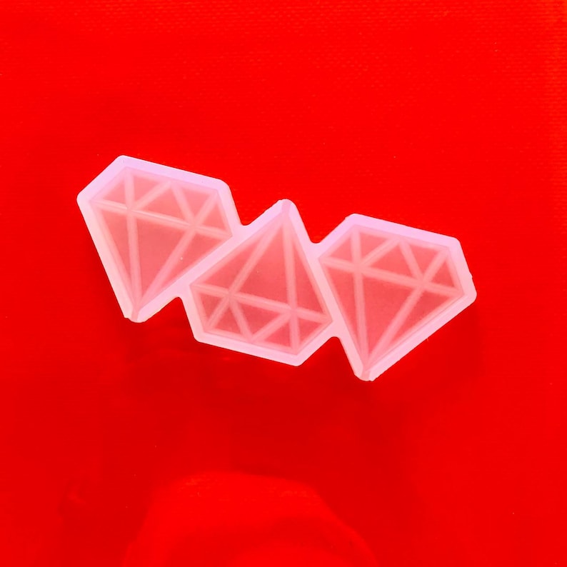 May include: Three clear diamond-shaped molds with geometric designs are arranged on a red background. The molds are made of a translucent material, and each diamond has multiple facets. The overall theme is related to crafting or jewelry making.