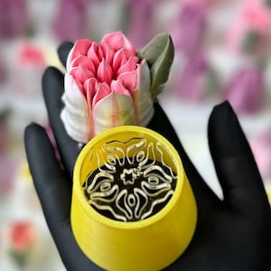 May include: A close-up of a floral design cake with pink and white petals and a green leaf. A yellow container with a decorative design is in the foreground. The background is blurred with pink and purple flowers.