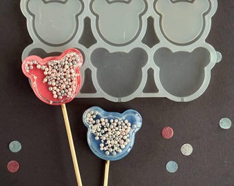 May include: A silicone mold with six bear-shaped cavities, next to two bear-shaped lollipops on wooden sticks. One lollipop is red with pink and white sprinkles, the other is blue with white sprinkles. Confetti dots are scattered around.