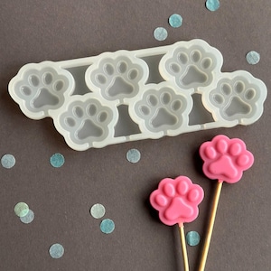 May include: A clear silicone mould with six paw prints and two pink paw print lollipops on wooden sticks. The paw prints are on a dark grey surface with scattered blue confetti.