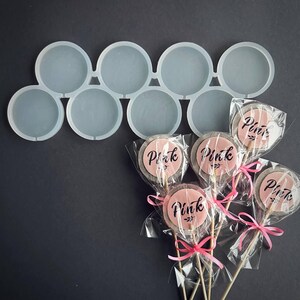 May include: A clear silicone mold with eight circular cavities, next to several wrapped lollipops. The lollipops have a pink center with the word "Pink" in black script, tied with pink ribbons.