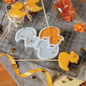 May include: A selection of handmade lollipops shaped like ducks and squirrels, presented on a wire rack. The lollipops are in shades of orange and yellow, some wrapped in clear cellophane. Gold beads and orange flower decorations are also included.
