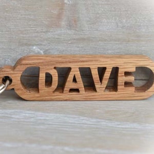 May include: A wooden keychain with the name "DAVE" cut out in capital letters. The keychain is made of light brown wood and has a silver metal keyring attached. The background is a light gray wooden surface.