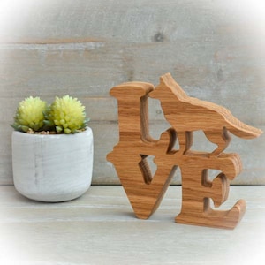 May include: A wooden "LOVE" sculpture with a dog silhouette on the "V". The light brown wood sculpture is on a light surface. A small white pot with two yellow-green succulents is to the left. The scene has a rustic feel.