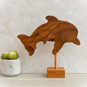 May include: A wooden dolphin sculpture featuring a larger dolphin with a smaller dolphin attached. The sculpture is made of stained wood and mounted on a wooden base. The dolphins are a medium brown color and have simple, carved details.