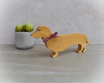 May include: A wooden dachshund dog ornament with a red and blue tartan ribbon. The dog is light brown and has a smooth finish. A small potted plant with green succulents is in the background.