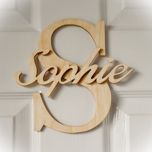 May include: A wooden letter 'S' with the name 'Sophie' in a cursive font, attached to the letter. The wooden sign is light brown and is mounted on a white door.