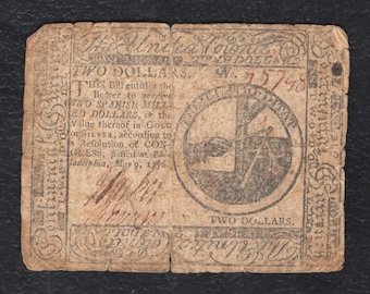 May include: A vintage two-dollar Continental currency bill from 1776, printed on aged tan paper with black ink. It features the text 'The United Colonies', 'Two Dollars', and a circular emblem depicting a hand holding a balance scale.