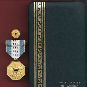 Puede incluir: Una medalla de oro con una cinta azul y blanca, un pequeño pin de oro y una caja negra con ribetes dorados. La caja tiene las palabras "United States of America" impresas en la parte delantera.