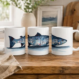 May include: Three white ceramic mugs featuring a fish design. The fish illustration is blue and white, with a geometric pattern. The mugs are on a wooden surface, with a neutral-coloured cloth in the foreground.