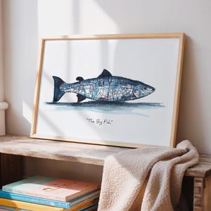 May include: Framed art print of a blue and grey fish illustration, with the text "The Big Fish". The artwork is in a light wooden frame, resting on a wooden shelf with books and a beige blanket.