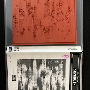 May include: A clear plastic package containing a set of two rubber stamps. The stamps are designed to create a dry brush effect. The package includes a stamp with a wood grain pattern and a stamp with a dry brush stroke pattern. The package is labeled "Drybrush Cling Stamp Set".