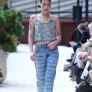 May include: A model on a runway wearing a light green patterned crop top and high-waisted, light blue jeans with a binary code design. The model is also wearing platform heels.