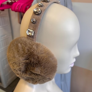 Puede incluir: Orejeras color topo con cubiertas de orejas de piel sintética y una diadema adornada con gemas claras y brillantes. Las orejeras se exhiben en una cabeza de maniquí, mostrando el diseño.