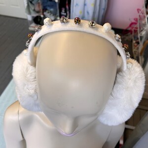 Puede incluir: Orejeras de piel sintética blanca con una diadema decorativa. La diadema está adornada con pequeños pedrería brillantes y adornos blancos en forma de calavera. Las orejeras son suaves y esponjosas, proporcionando calidez y estilo.