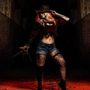 Pode incluir: Uma mulher com chapéu castanho e top listrado vermelho e preto, segurando uma luva com lâminas. Ela usa shorts jeans, meias de rede e botas pretas. A cena é fracamente iluminada com tons vermelhos e laranja, e folhas espalhadas.