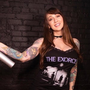 Pode incluir: Uma mulher vestindo um vestido preto com uma estampa gráfica branca do filme "O Exorcista". Ela tem cabelo castanho longo e está usando um colar preto de choker. Ela tem tatuagens nos braços e nas pernas.