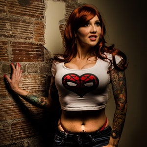 Pode incluir: Uma mulher com cabelo ruivo está vestindo um top branco curto com um desenho de coração vermelho e jeans azul. O coração tem um padrão de teia de aranha e um rosto de aranha branco. Ela tem tatuagens nos braços.