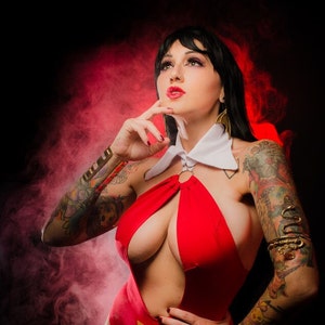 Pode incluir: Uma mulher com um traje vermelho de uma peça, de decote profundo, com um colarinho branco e uma pulseira dourada. Ela tem cabelo comprido e preto e está usando batom vermelho. Ela está em pé em frente a um fundo vermelho e preto fumegante.