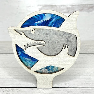 May include: A wooden shark-shaped decoration with a white frame. The shark's body is gray, and the background features blue wave patterns. The design includes cut-out sections revealing the wave pattern, creating a layered effect. The decoration has a stand.