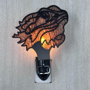 May include: A decorative night light featuring a dragon head design. The dragon head is black with a wood-grain pattern, and the light emits a warm orange glow. The night light plugs directly into a wall outlet.