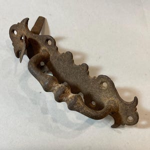 May include: A rusty, dark brown metal drawer pull featuring an ornate, elongated backplate with scalloped edges and four circular screw holes. The handle itself has a textured, segmented design and is attached to the backplate with two hinges.