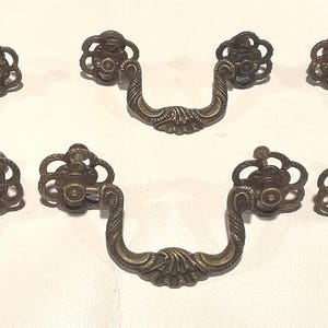 May include: Set of seven antique bronze drawer pulls. Each pull features a curved handle with ornate floral detailing and matching circular backplates. The hardware is designed for furniture restoration or adding vintage charm to cabinets and drawers.