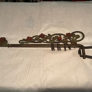 May include: An antique bronze curtain rod with decorative floral accents. The rod features red roses and green leaves, with hooks for hanging curtains. The rod is supported by a bracket on the right side.