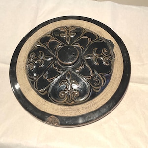 May include: A circular ceramic lid featuring a glossy black glaze on a raised, embossed floral pattern. The centre is surrounded by a wide, unglazed beige border with a textured finish. The outer edge is finished in a smooth, dark black glaze.