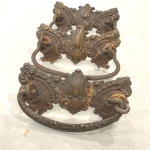 May include: Three antique bronze drawer pulls with ornate floral and scrollwork designs. Each pull features a curved handle and decorative backplate with intricate detailing. The pulls are aged with a patina finish.