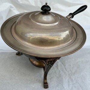 May include: Silver plated chafing dish with a black handle and a round, ornate base with three legs. The dish has a lid with a black knob.