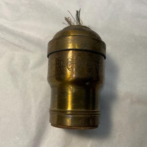 May include: A cylindrical brass lamp socket featuring a weathered metallic finish and a visible wick at the top. The body displays a circular logo with the text Rubber and a series of horizontal grooves near the base, showing signs of age and use.