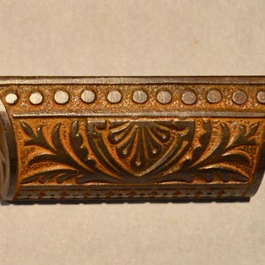 May include: A bronze-toned metal drawer pull featuring a horizontal row of circular indentations along the top edge and a central embossed floral leaf pattern. The hardware includes two mounting holes on the flat side extensions for attachment.