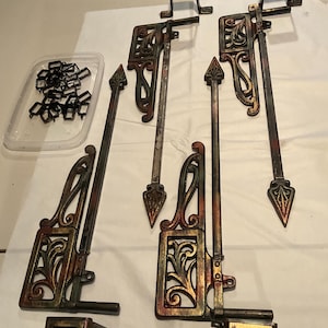 May include: An assortment of antique metal hardware pieces, including ornate brackets, spear-topped rods, and small rectangular components. The pieces have a distressed finish with shades of bronze, copper, and red, suggesting an aged appearance. A plastic container holds small, black metal connectors.