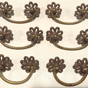 May include: Set of six antique brass drawer pulls. Each pull features a floral design on the ends and a curved handle. The brass has a warm, aged patina, adding to the vintage aesthetic. Ideal for restoring furniture.