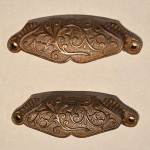 May include: Two bronze-toned metal drawer pulls featuring an embossed floral scrollwork pattern across the center. Each handle has a curved, elongated shape with a textured surface and two circular screw holes on the outer ends for mounting.