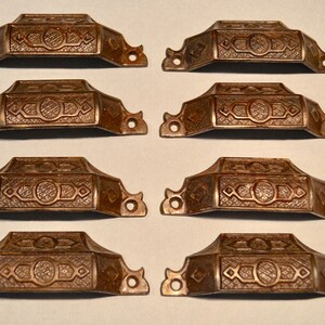 May include: A set of eight matching bronze-toned metal drawer pulls arranged in two columns. Each pull features a rectangular central panel with an embossed cross-hatched pattern, flanked by two screw holes on flat, decorative side extensions.