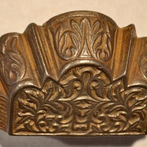 May include: A bronze-toned metal drawer pull featuring an ornate, raised relief design with intricate floral and leaf motifs. The hardware has a central arched section with a decorative pattern and two side mounting points with circular screw holes.