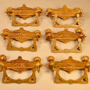 May include: Six antique brass drawer pulls. Each pull has a decorative rectangular plate with an ornate design and a curved handle. The pulls are a warm gold color and are arranged in a grid pattern.