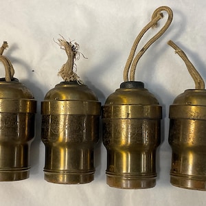 May include: Four antique brass light bulb sockets with cylindrical bodies and weathered finishes. Each socket features a top-mounted beige fabric cord loop for hanging and embossed text on the side reading 250W 250V.