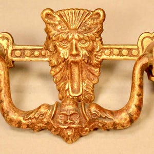 May include: Ornate gold-colored drawer pull with a central face design and curved handles. The face has a beard and a crown-like feature. The pull has decorative elements and a vintage aesthetic, suitable for antique furniture.