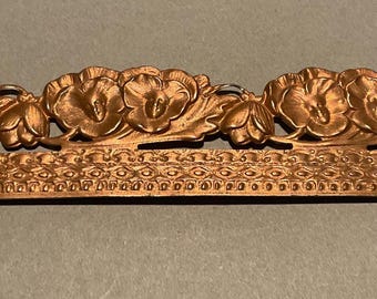 May include: A copper-colored decorative metal piece featuring a floral design. The top portion showcases stylized flowers, while the bottom has a repeating pattern. The piece is likely for decorative purposes, possibly for furniture or crafts.