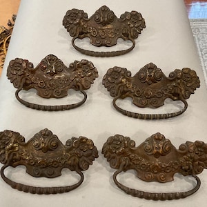 May include: Five antique bronze drawer pulls with ornate floral and scrollwork designs. Each pull has a curved handle and a decorative backplate. The pulls are a warm, aged bronze color, suggesting vintage hardware.
