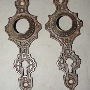 May include: Two antique, ornate metal escutcheons with keyholes. The escutcheons are a tarnished silver color, featuring a decorative design with a circular top, a keyhole, and a decorative bottom. The design is symmetrical and detailed.
