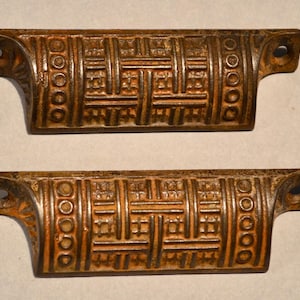 May include: Two matching bronze-toned metal drawer pulls featuring a raised, textured basket-weave pattern across the center and a series of circular indentations on the sides. Each handle has two mounting holes for attachment to furniture.