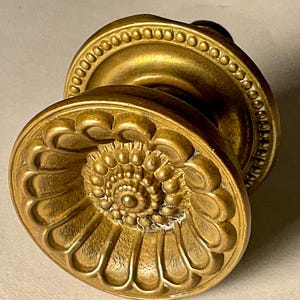 May include: A vintage brass door knob with an ornate floral design. The knob features a detailed, raised pattern with a central spiral and a beaded edge. The brass has a warm, aged patina.