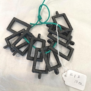 May include: A collection of dark gray, rectangular metal charms with a textured finish, strung together with green and white cord. Each charm has a small loop at the bottom. A white tag with the text "#1 A 10 ea" is attached.